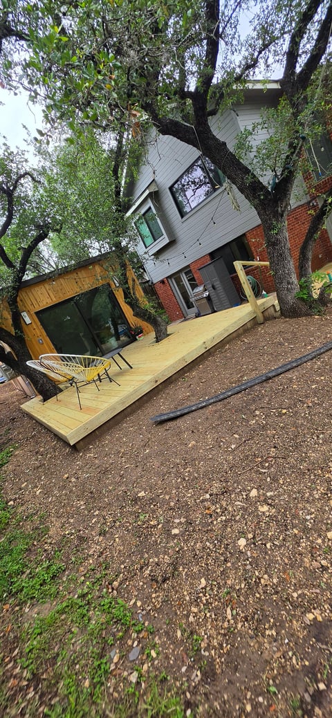 Modern tiny house with wooden deck nestled under large tree, featuring white upper level and brick base with adjacent wooden structure
