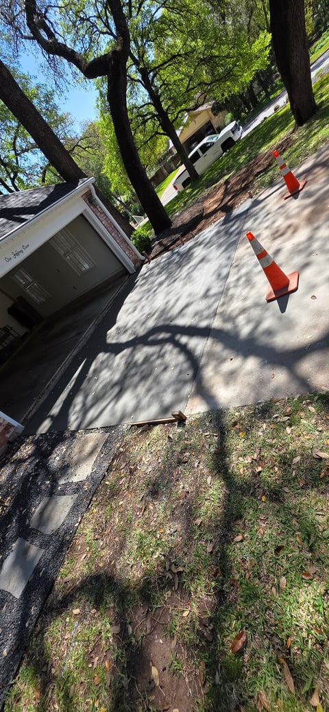 Residential driveway with orange traffic cones, large trees providing shade, and a building visible in the background