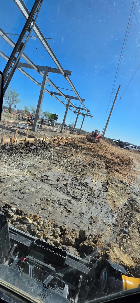 Construction site with metal structure frame under blue sky, dirt pile and heavy machinery visible